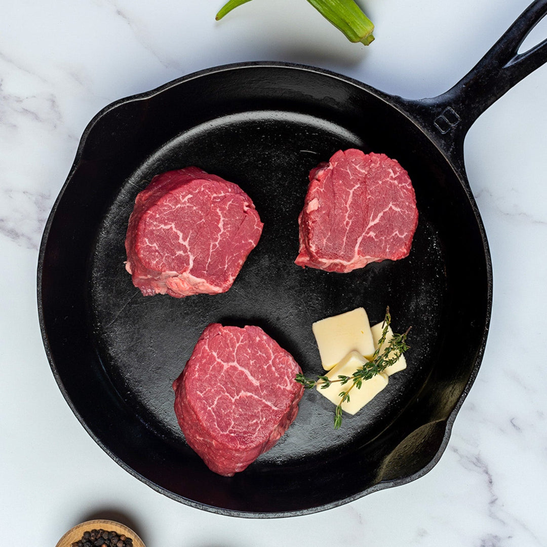 Three raw steaks in a cast iron skillet with butter and herbs on a marble surface.